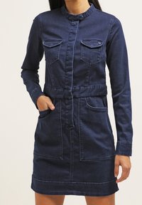 Tummansininen farkkumekko, jossa mandariinikaulus, pitkät hihat, kaksi rintataskua ja vyötäröllä oleva vyö. Pehmeä koostumus.