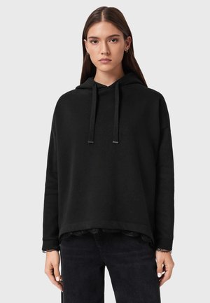 Schwarzer Hoodie mit lockerem Schnitt, Kapuze mit Kordelzug und strukturiertem Saum. Der Stoff ist weich und wird über einem gemusterten schwarzen Oberteil getragen.