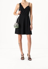 Nainen pukeutuneena hihattomaan mustaan kietaisumekkoon, mustiin nauhakahvoihin ja kantaen pientä hopeista ketjuhihnalla varustettua käsilaukkua. Jaloissa näkyviä tatuointeja.