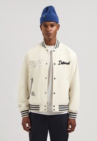 Hvid bomberjakke med marineblå stribede ribmanchetter og krave, broderede detaljer og quiltet tekstur. Båret over en grå skjorte.