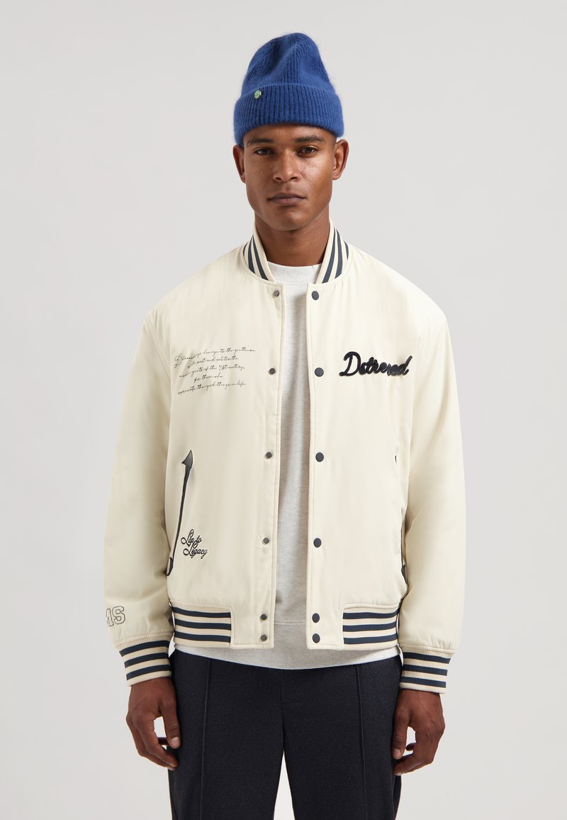 Hvid bomberjakke med marineblå stribede ribmanchetter og krave, broderede detaljer og quiltet tekstur. Båret over en grå skjorte.