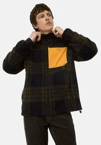 Fleecejacke in dunkelgrün und schwarzem Karomuster mit einer orangefarbenen Brusttasche. Sie verfügt über einen Reißverschluss, einen hohen Kragen und Seitentaschen.