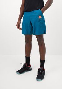 Turquoise athletic shorts with side pockets and an orange logo; paired with black shoes featuring red accents and black socks.