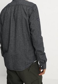 Camisa de color gris oscuro con estampado, mangas largas, puños con botones y dobladillo curvo. Cuenta con un pequeño parche con logo en la manga izquierda.