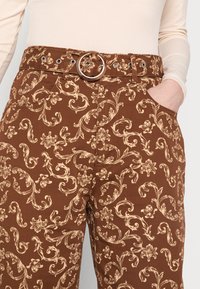 Calças castanhas com padrão floral, apresentando um cinto correspondente com uma fivela metálica redonda e bolsos laterais. Textura de mistura de algodão visível.