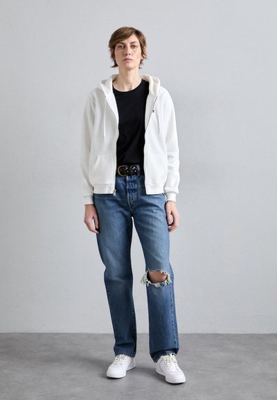 Haut à zip blanc porté sur un t-shirt noir, associé à un jean en denim bleu avec un genou déchiré. Porté avec des baskets blanches et une ceinture noire.