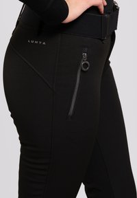 Schwarze Sporthosen mit strukturiertem Finish, ausgestattet mit einer Reißverschlusstasche und einem breiten Bund. Das Markenzeichen ist am Hüftbereich sichtbar.