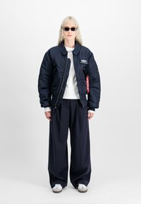 Person som har på seg svarte solbriller, en stor marineblå bomberjakke, hvit skjorte, vide marineblå bukser og hvite joggesko, som står mot en ensfarget bakgrunn.