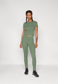 Olivově zelený crop top a legíny s vysokým pasem vyrobené z elastického materiálu. Má hladkou texturu a těsně padnoucí design. Černé sportovní boty.