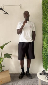 White t-shirt with a logo, black shorts, and black shoes. Standing against a textured wall with green foliage and potted plant nearby.