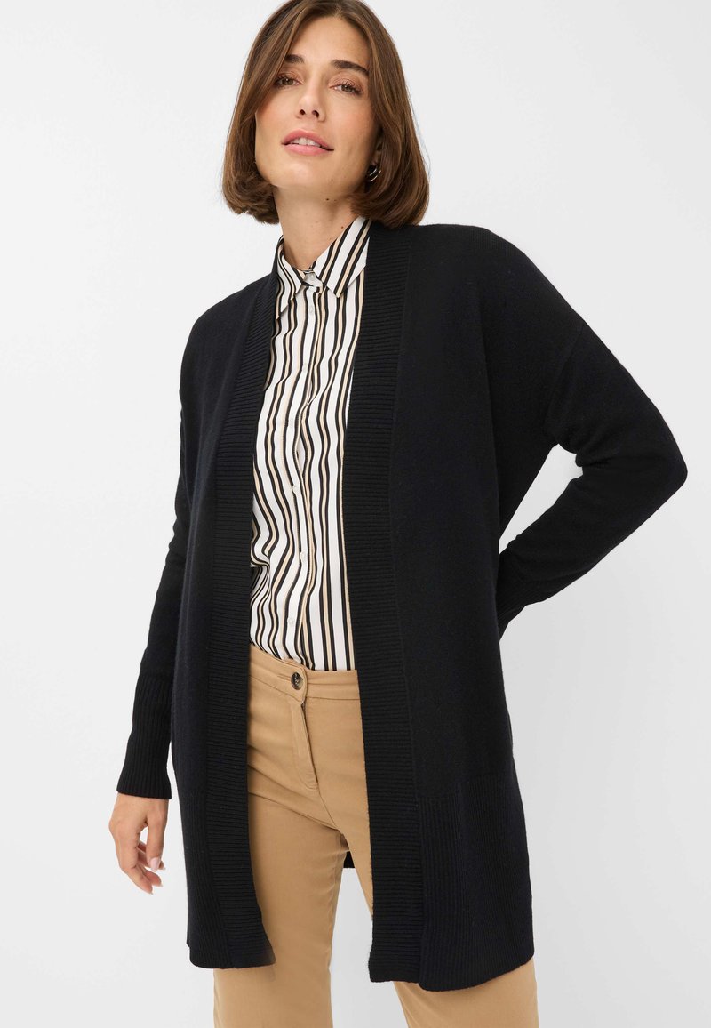 Schwarze gestrickte Strickjacke mit offenem Vorderteil und gerippten Besätzen, kombiniert mit einem gestreiften Hemd. Beigefarbene Hose mit einem Knopfdetail. Schlichtes, lockeres Design.