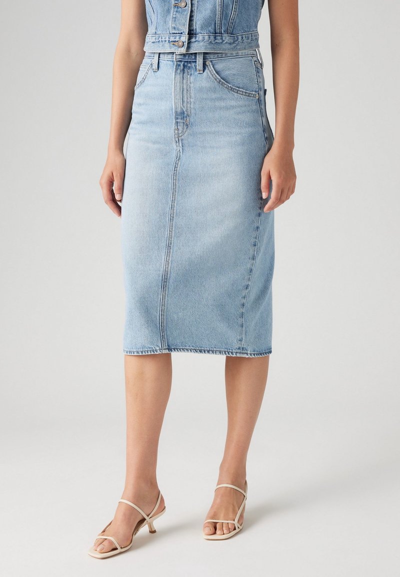 Femme portant une jupe crayon en jean bleu clair et un gilet en jean sans manches assorti, associée à des sandales à talons beiges avec des brides.