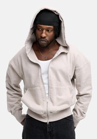 Light beige zip-up hoodie made from soft fabric, featuring a large hood, front pockets, and ribbed cuffs. Worn over a white tank top.