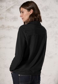 Maglietta nera a maniche lunghe con colletto e dettaglio sullo yoke posteriore, realizzata in materiale morbido; indossata con jeans scuri.