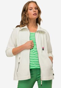 Giacca bianca con zip e collo alto, dotata di due tasche frontali, bordi grigi e un cordino. Indossata sopra una maglietta a righe verdi e pantaloni verdi.