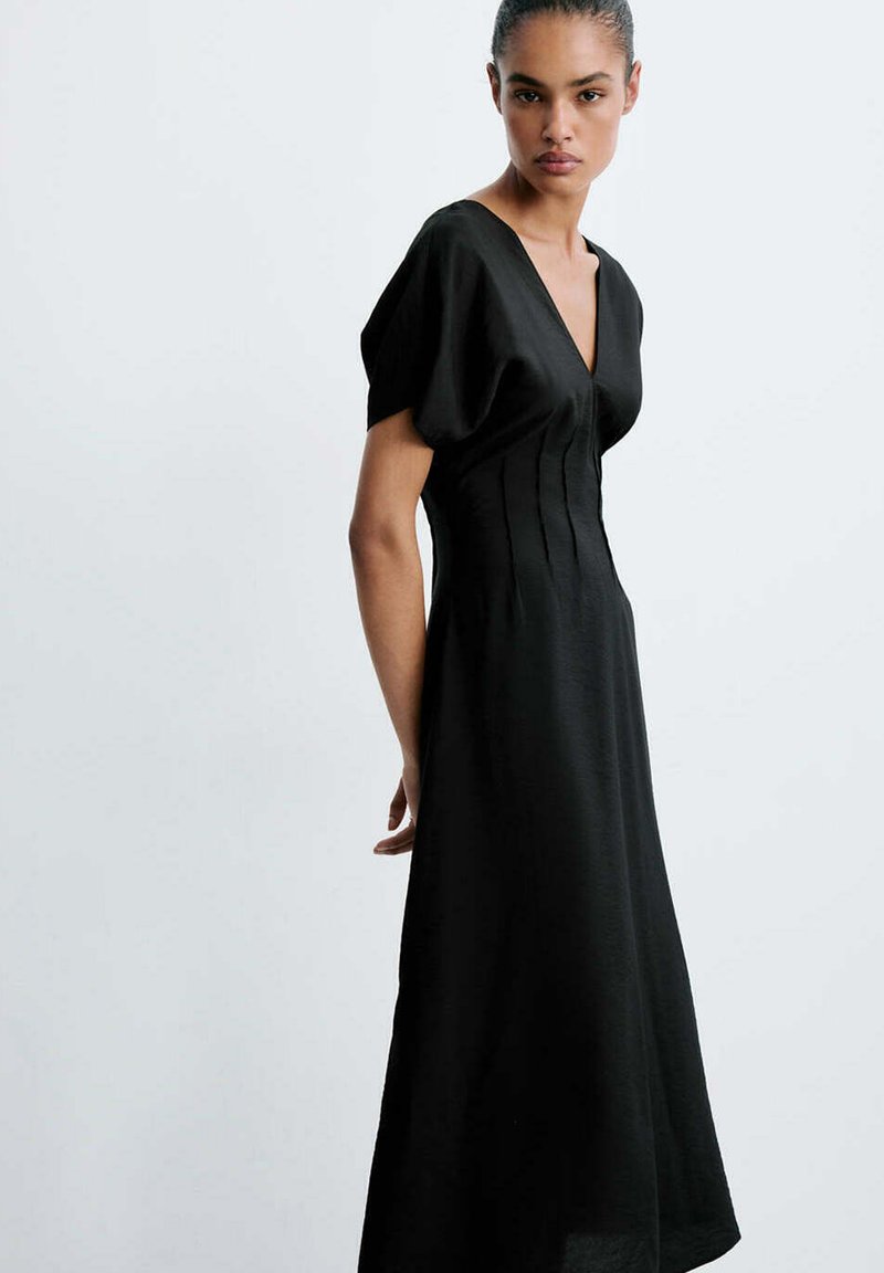 Femme aux cheveux tirés en arrière, portant une robe noire à manches courtes et col en V, avec une taille ajustée, debout devant un fond clair uni.