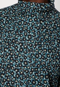 Haut à imprimé floral avec col montant. Arrière-plan principalement bleu canard avec des fleurs noires et des accents beiges. Texture de tissu douce.