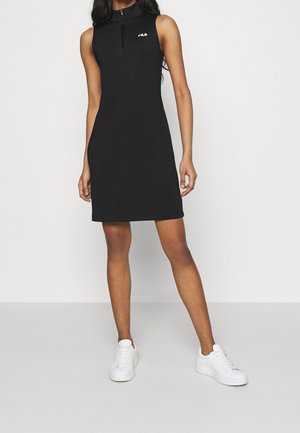 Femme portant une robe noire sans manches zippée avec le logo Fila et des baskets blanches, debout devant un fond clair uni.