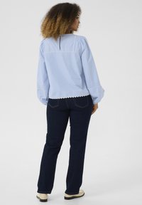 Camicia azzurra a maniche lunghe con maniche a sbuffo, rifiniture in pizzo, apertura a chiave sul retro, abbinata a jeans flare di denim scuro e scarpe chiare.