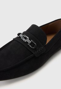 Czarne mokasyny zamszowe o płaskim kształcie, zaokrąglonym nosku i ozdobnym srebrnym łańcuchem z przodu. Teksturowana powierzchnia z detalami szwów.