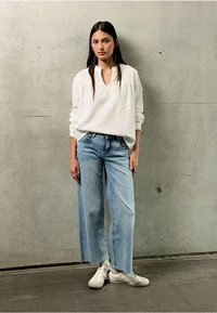 Blusa bianca a maniche lunghe con texture e scollo a V, abbinata a jeans a gamba larga blu chiaro e sneakers bianche. Sfondo in calcestruzzo grigio.