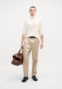 Pantalons beiges, pull à col roulé crème, ceinture noire et chaussures noires ; tenant un sac en cuir marron avec une texture douce et une forme structurée.