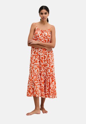 Femme debout pieds nus, bras croisés, portant une robe midi sans manches à fleurs orange et blanche avec un ourlet volanté.