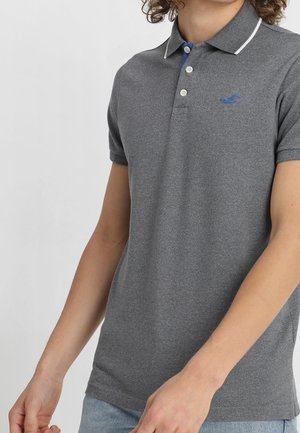 Jeune personne portant un polo gris cintré avec une bordure blanche sur le col et un logo bleu sur la poitrine.