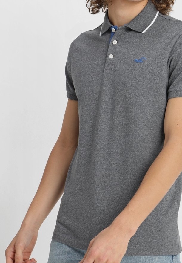 Jeune personne portant un polo gris cintré avec une bordure blanche sur le col et un logo bleu sur la poitrine.