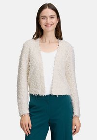 Cardigan en tricot épais couleur crème avec un tissu texturé, devant ouvert et bords effilochés. Porté sur un haut blanc et associé à un pantalon teal.