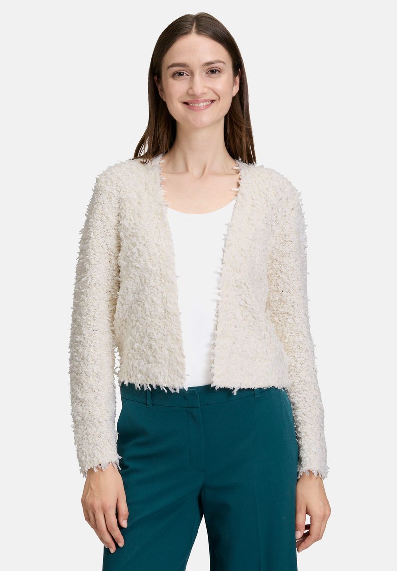 Cardigan en tricot épais couleur crème avec un tissu texturé, devant ouvert et bords effilochés. Porté sur un haut blanc et associé à un pantalon teal.