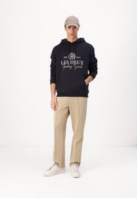 Felpa navy con testo e logo grafico bianchi, tasca anteriore, abbinata a pantaloni beige chiaro e sneaker bianche. Il modello indossa un cappellino beige.
