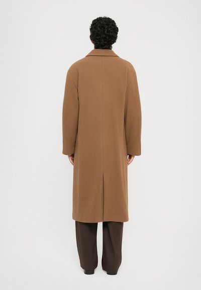 Man facing away wearing a long camel-colored coat, dark brown pants, and black shoes against a plain white background.