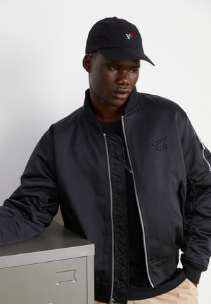 Black bomber jacket with zipper, ribbed cuffs, Nike logo, worn over a black shirt. Black cap with rose embroidery. Metal filing cabinet nearby.