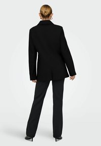 Musta räätälöity bleiseri terävillä läpillä ja yksittäisellä halkiolla, yhdistettynä istuviin mustiin housuihin ja korkokenkiin, esittelee virtaviivaista siluettia.