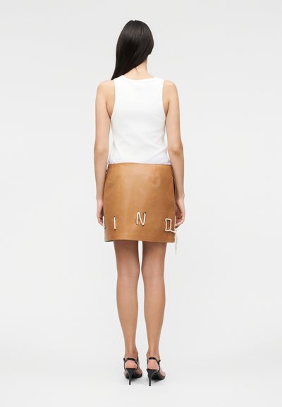 Γυναίκα με μακριά μαύρα μαλλιά που φοράει λευκή αμάνικη μπλούζα και καφέ δερμάτινη μίνι φούστα με λευκά δαντελένια γράμματα στην πλάτη.