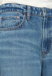 Jeans de mezclilla azul claro con herrajes en tono cobre, diseño clásico de cinco bolsillos y patrones de costura visibles. Tejido texturizado con un acabado desgastado.