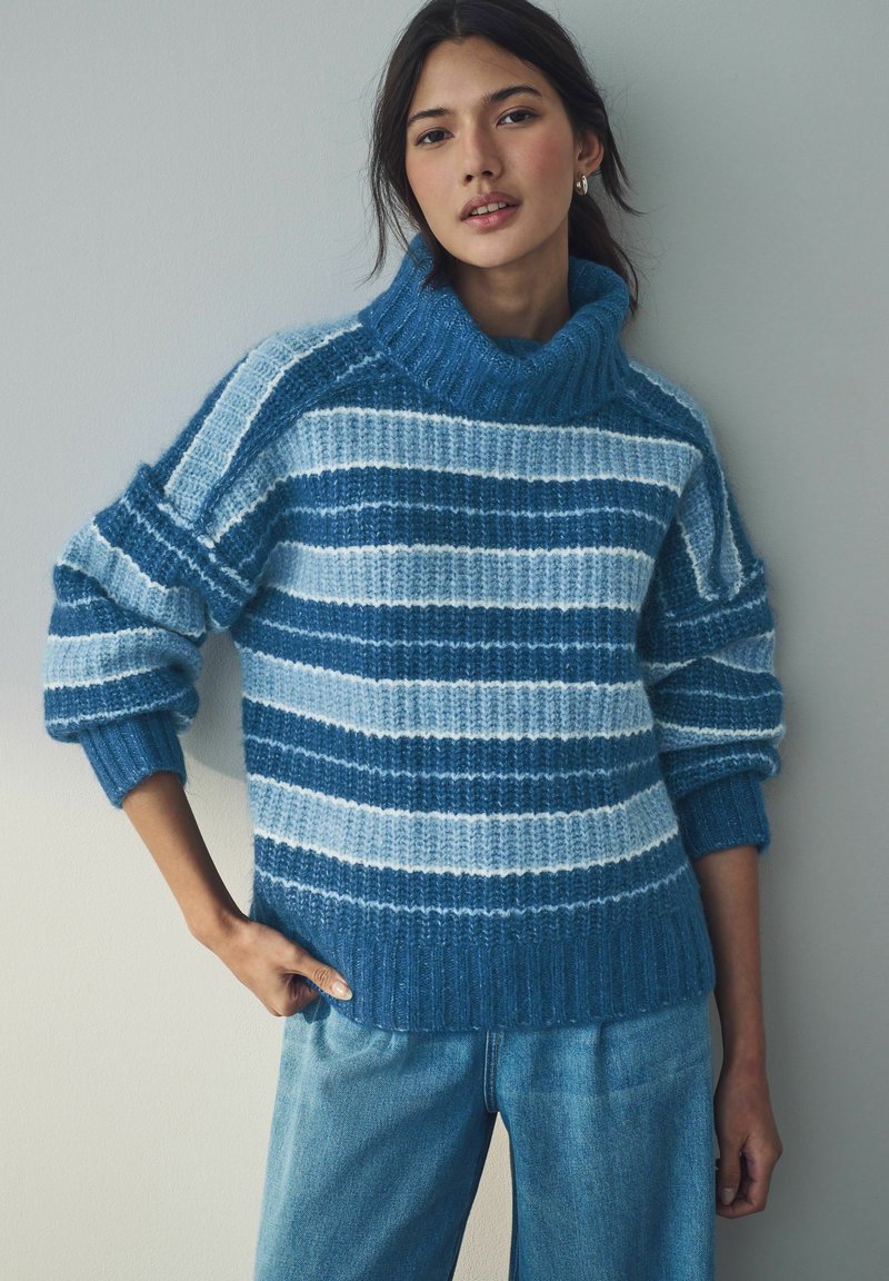 Junge Frau in einem blau- und hellblau gestreiften, gestrickten Rollkragenpullover und hellblauen Jeans, steht vor einer einfarbigen Wand.