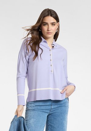 Femme aux longs cheveux bruns portant un pull boutonné violet clair et un jean à motifs floraux bleus, tenant une veste en jean dans une main.