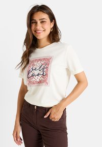 Camiseta de algodón blanca con un diseño de borde paisley rojo y texto "amor propio". Manga corta, corte relajado, combinada con pantalones marrones oscuros.