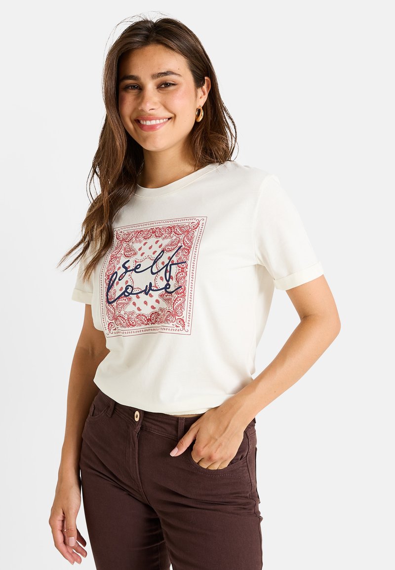 Camiseta de algodón blanca con un diseño de borde paisley rojo y texto "amor propio". Manga corta, corte relajado, combinada con pantalones marrones oscuros.