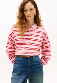 Πόλο μπλούζα με μακριά μανίκια σε ανοιχτό ροζ χρώμα με κόκκινες ρίγες, με γιακά και κουμπιά. Φοριέται με τζιν παντελόνι μπλε ντένιμ και καφέ ζώνη.