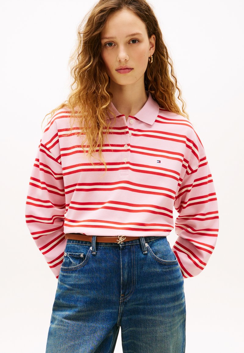Πόλο μπλούζα με μακριά μανίκια σε ανοιχτό ροζ χρώμα με κόκκινες ρίγες, με γιακά και κουμπιά. Φοριέται με τζιν παντελόνι μπλε ντένιμ και καφέ ζώνη.