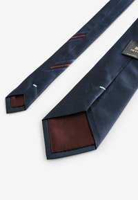 Mørkeblå silkebind med en tekstureret overflade, diagonale bordeaux- og blå striber samt en ensfarvet vinrød underside. Påsyet label synlig.