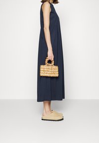 Navy sinivuorinen mekko, joka ulottuu nilkkoihin, yhdistettynä pieneen, vaaleanruskeasta ja beigeistä sävyistä kudottuun laukkuun. Beige liukuhihnat.