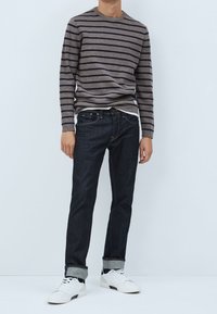 Camicia a maniche lunghe a righe marrone e blu navy sopra uno strato bianco, abbinata a jeans in denim scuro e sneakers bianche. Vestibilità casual, con i polsini arrotolati.