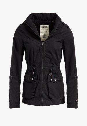 Personne portant une veste noire, une chemise grise, un jean bleu déchiré et des baskets blanches, debout sur un fond uni.