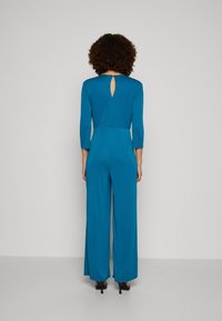 Jumpsuit turquoise avec des manches longues et un détail en clé à l'arrière. Fabriqué en tissu doux et lisse. Design à jambe large et taille cintrée.