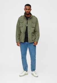 Oliven grøn militærjakke med fire forlommer, krave og knaplukning, parret med blå jeans og hvide sneakers.