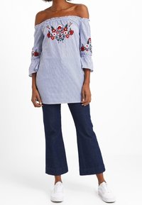 Sininen ja valkoinen raidallinen off-the-shoulder-paita, jossa on kukka-ompelukset hihoissa, yhdistettynä tummiin farkkulegginseihin ja valkoisiin lenkkareihin.